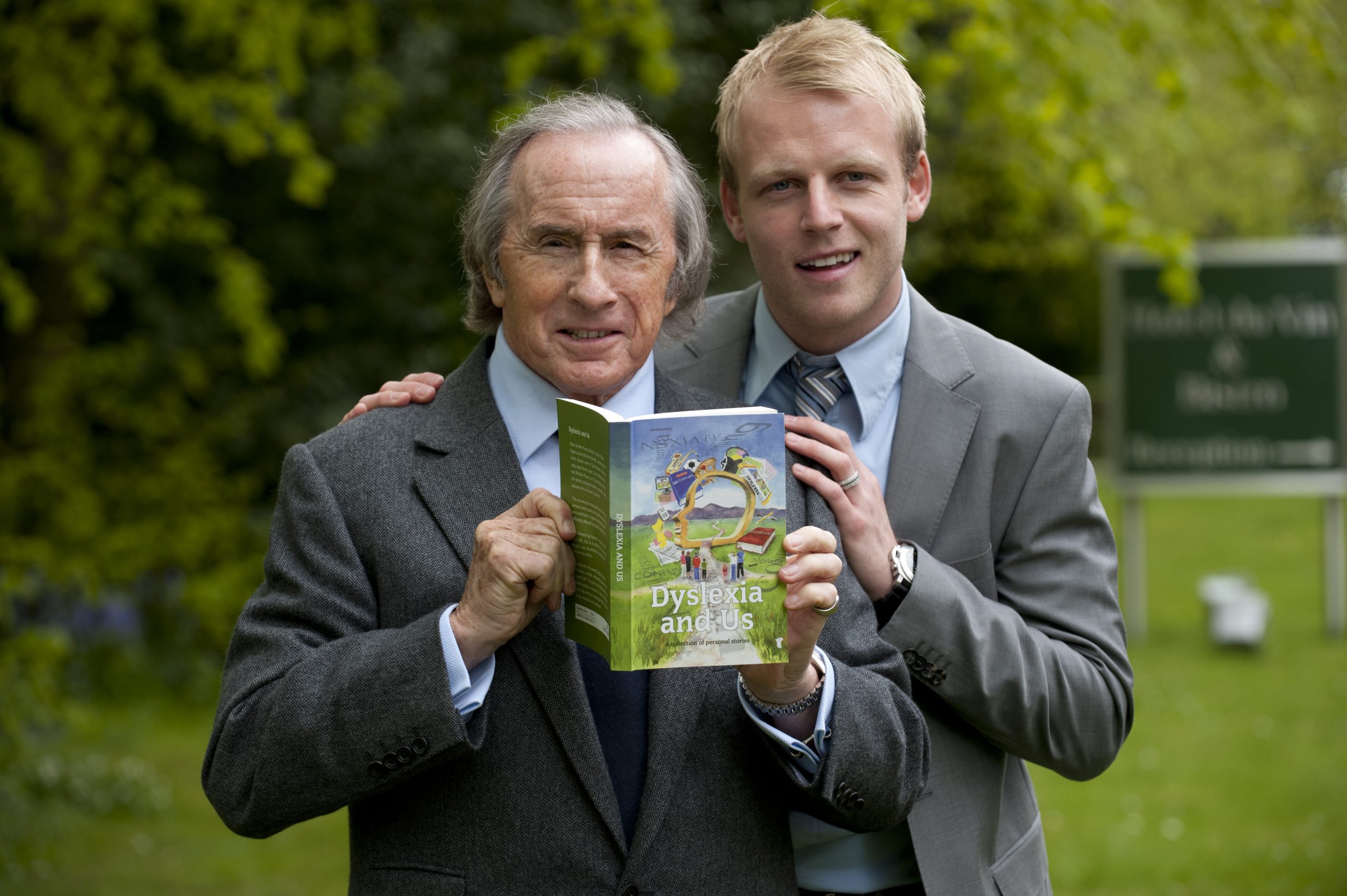 Celebritis Sir Jackie Stewart and Steven Naismith hold a copy of Dysleixa and Us between them; Steven's arm is placed around Jackie's shoulder and they both smile.