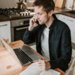 A man working at his kitchen table desk speaks on the telephone.