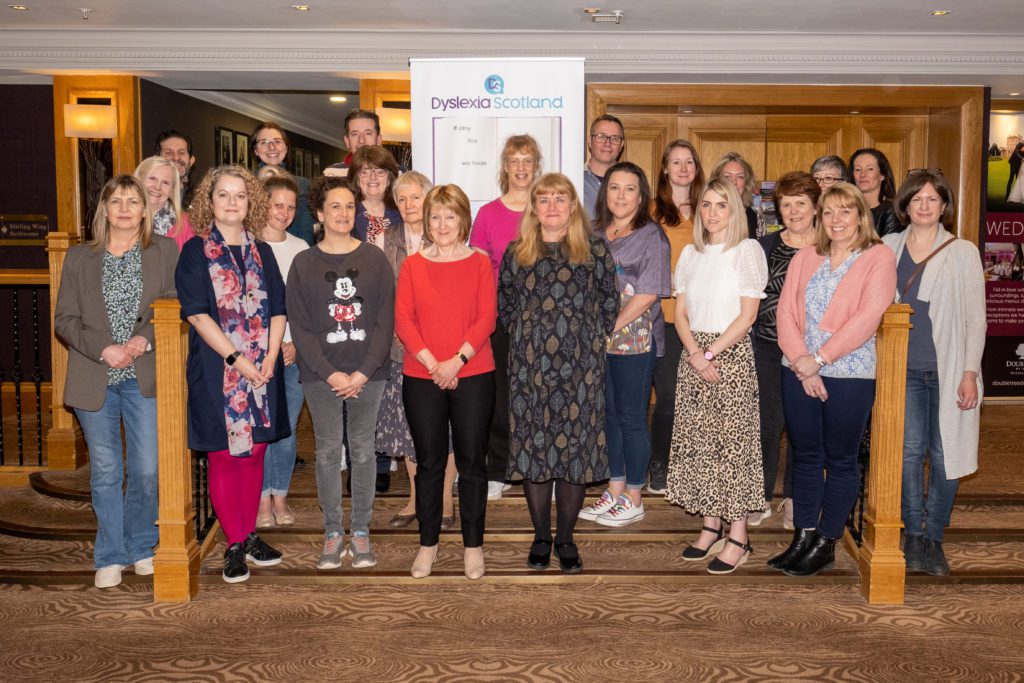 A large froup of Dyslexia Scotland local branch volunteers stand together in front of a Dyslexia Scotland pop up banner.