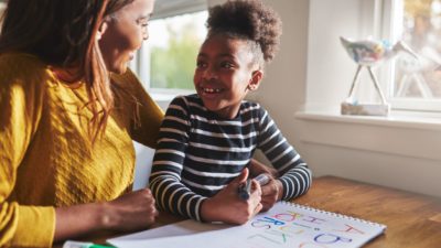 A mother puts her arm around her young daughter at the kitchen table. The daughter is drawing. They smile at one another.