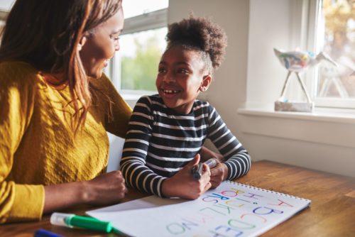 A mother puts her arm around her young daughter at the kitchen table. The daughter is drawing. They smile at one another.