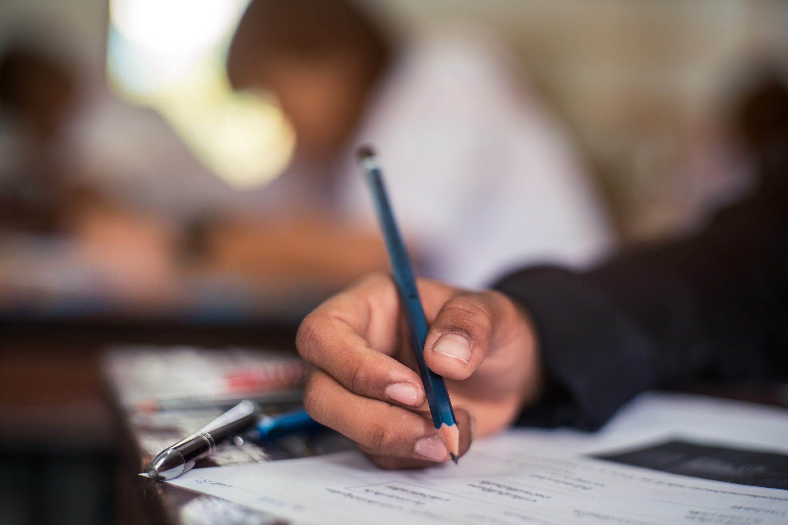 Close up of a hand holding a pencil. A blurred background shows a person in a white shirt leaned over at a desk.