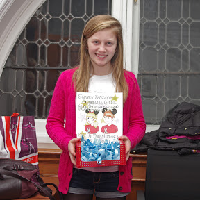 A young girl with blonde hair, wearing a bright pink cardigan, holds a box of blue awareness ribbons.