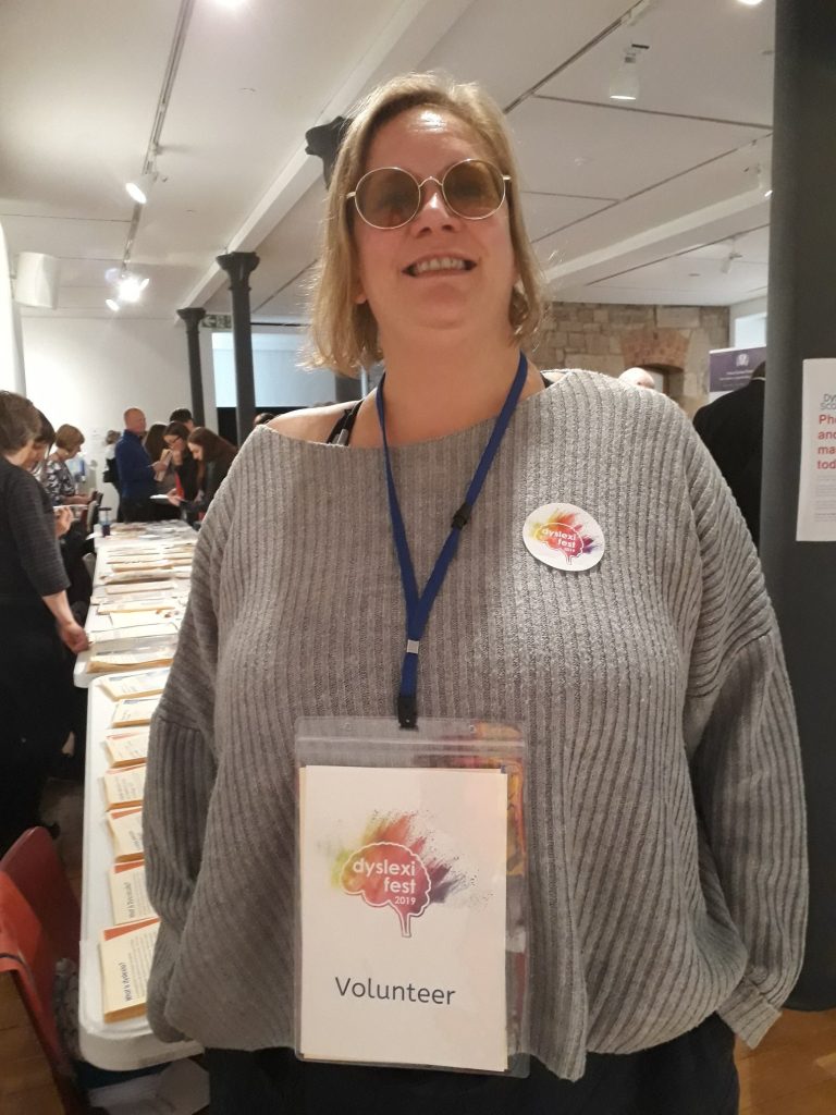 An event volunteer in the foreground of a busy room lined with stalls smiles at the camera, she is wearing a lanyard that says 'volunteer' on it.