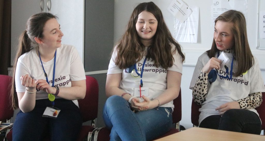 Three young people sat in a row engaging one another and smiling. They are each wearing a tshier that says 'Dyslexia Unwrapped' and brightly coloured volunteer lanyards.