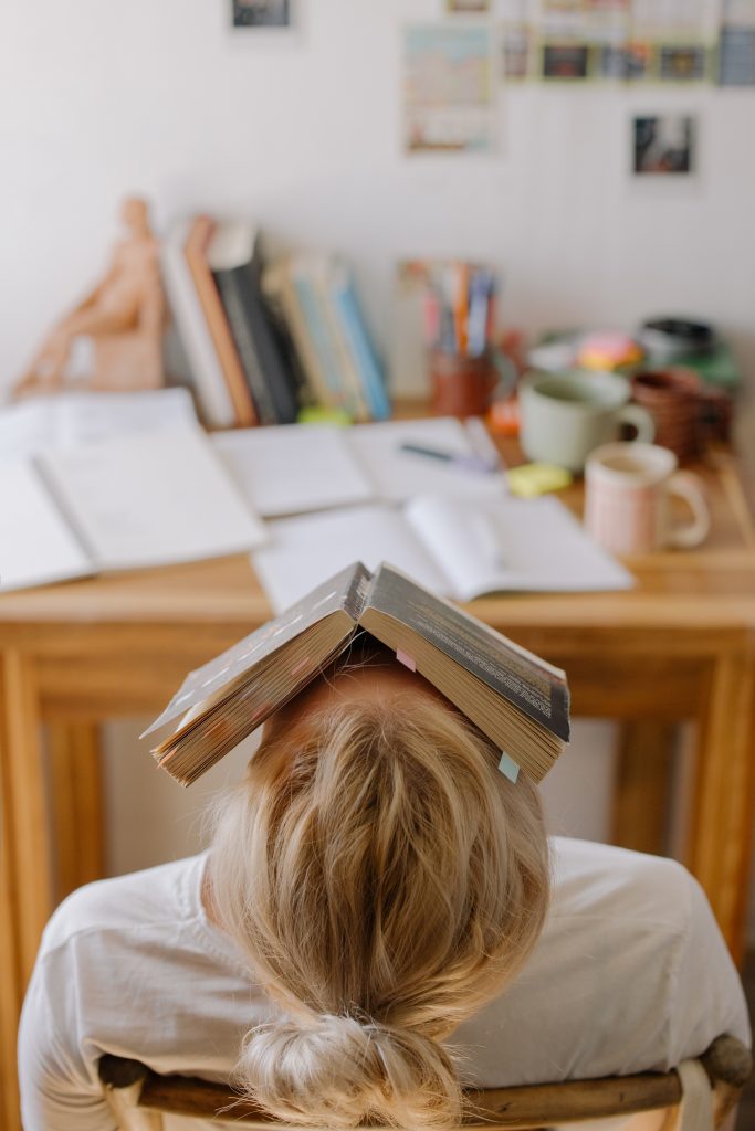 A woman leans back in her chair with a hardback book across her face. The desk in front of her is covered in study materials.