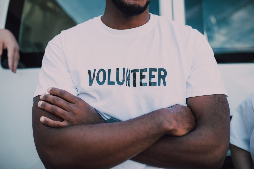 A close up of the chest and folded arms of a man. His white t-shirt has the word VOLUNTEER in black print.