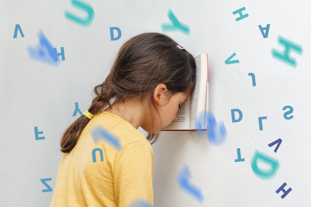 A girl leans face into a wall, her eyes closed, a book between her head and the wall. Blue and green letters fly randomly around her.