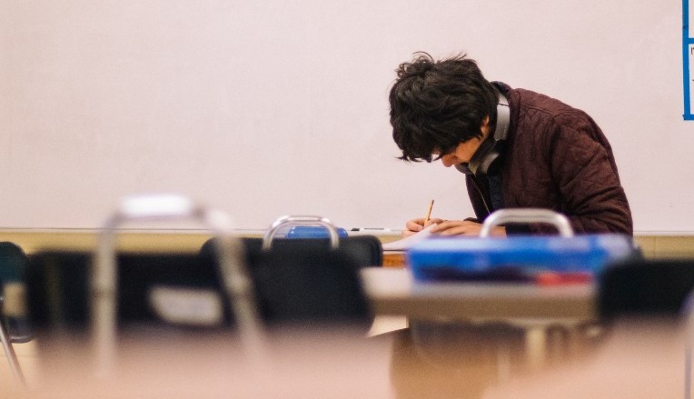 A young person in school uniform hunches over a desk, writing.