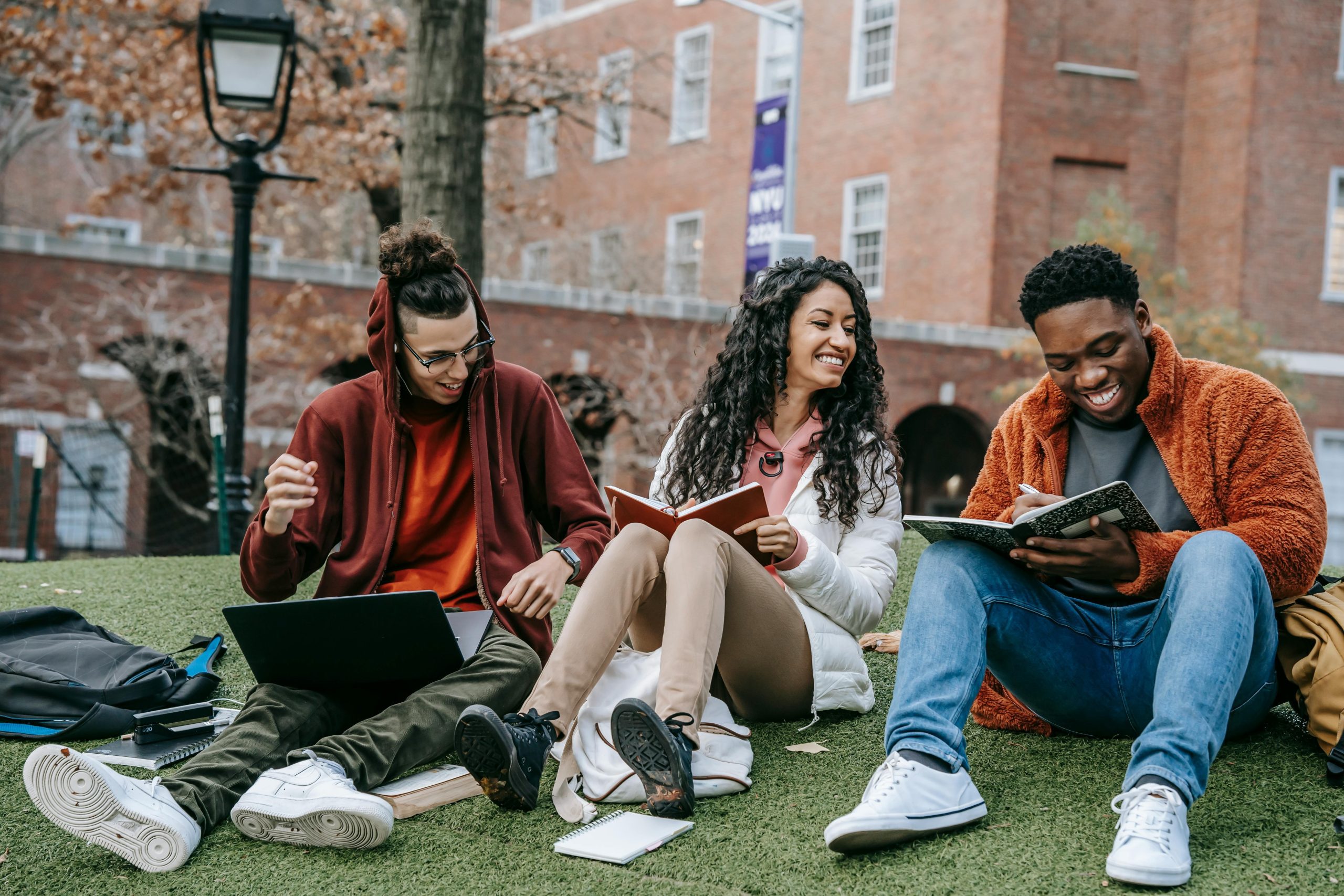 Three young people sit on the grass outside of their learning establishment, with rucksacks and books.