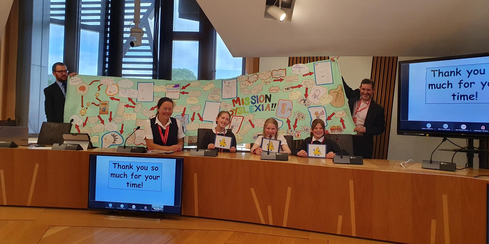 Oliver Mundell MSP and Paul McNeill, Convener of the CPG on Dyslexia in the Scottish Parliament hold up an enormous banner of children's work behind three young people and their teacher in a committee room.