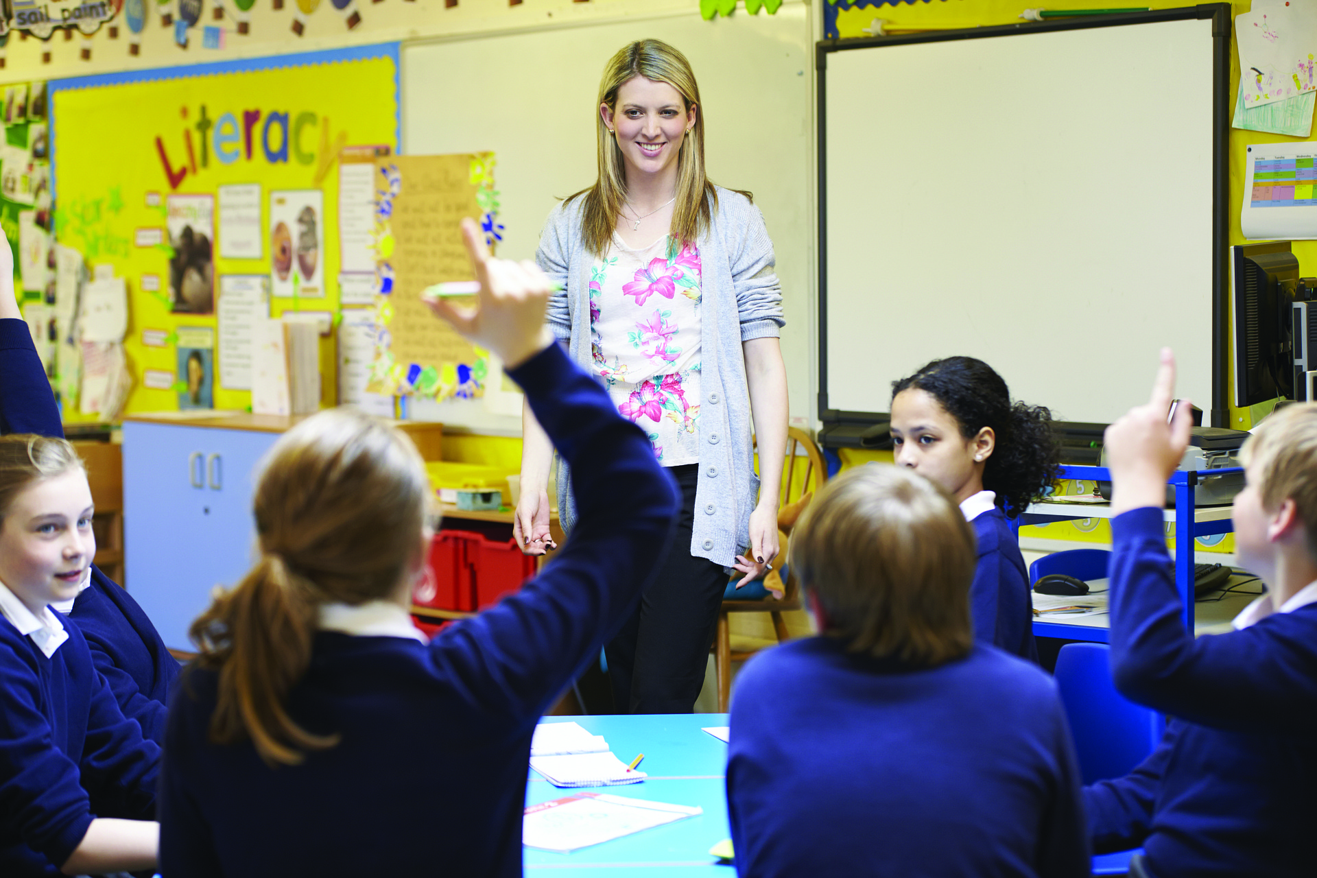 A learner in the classroom raises their hand to ask a question. A teacher smiles in front of the class.