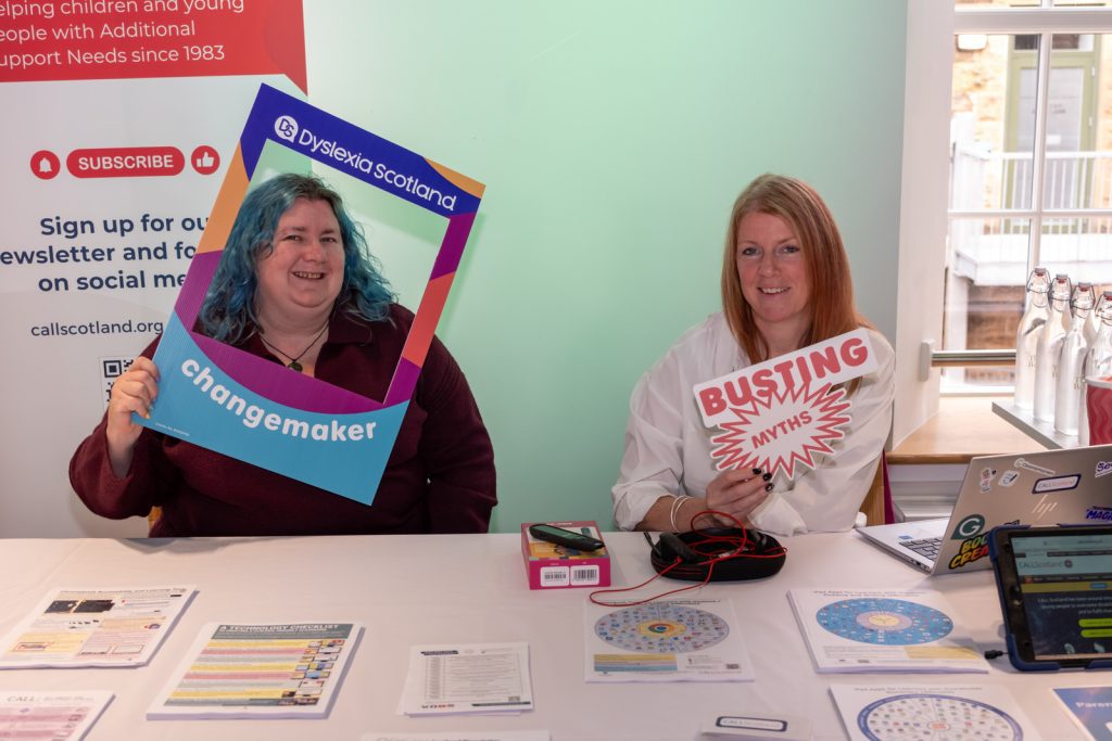 Smiling women at an information stall hold Dyslexia Scotland and Buting Myths signs