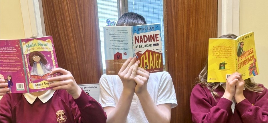Three children with their faces covered by the Gaelic books they are reading intently
