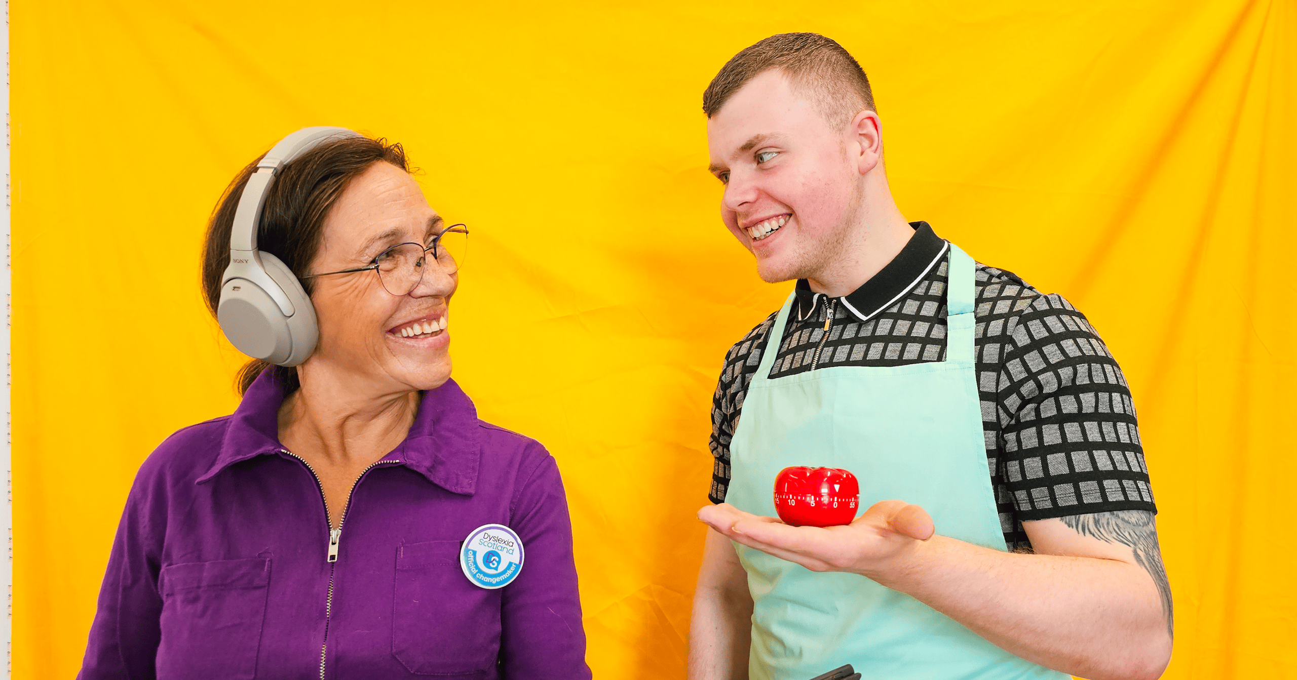 A woman wearing noise cancelling headphones smiles at a man who holds a pomodoro timer